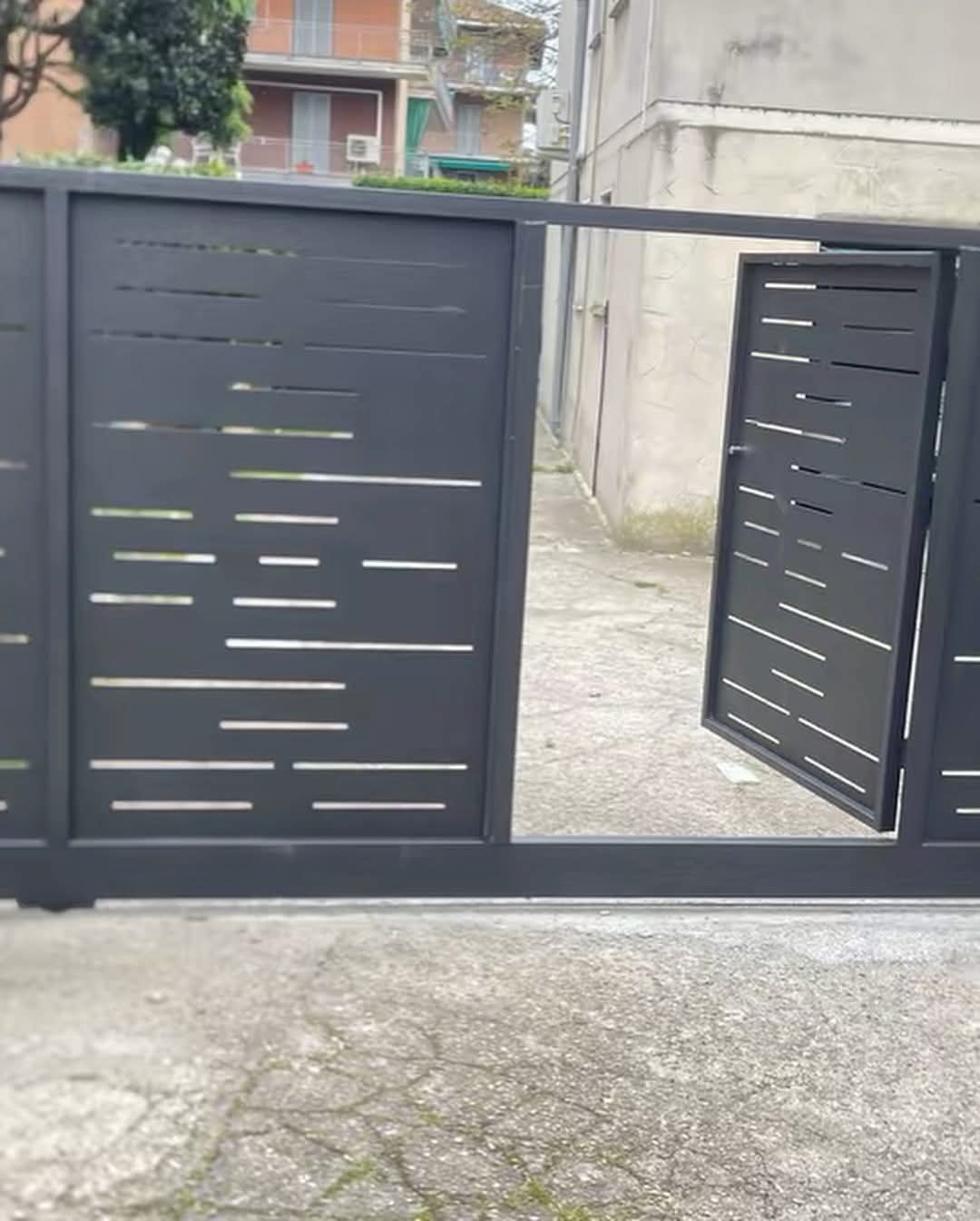 Cancello in metallo grigio scuro, parzialmente aperto, con ritagli orizzontali e irregolari. Terreno in cemento e sfondo dell'edificio.
