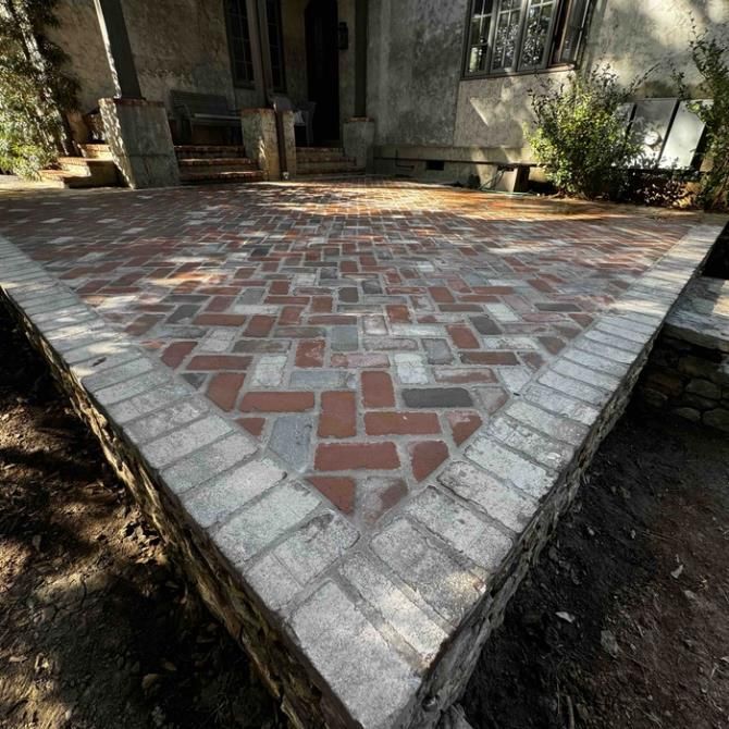 Person laying gray paving stones in a sandy area, using gloves, next to bags and tools.