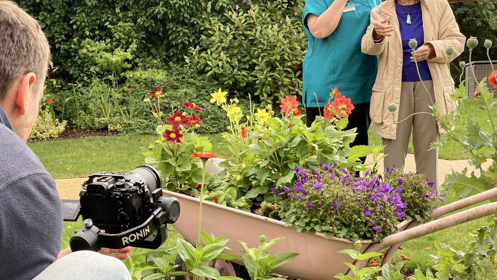 A man is taking a picture of a woman in a wheelbarrow in a garden.