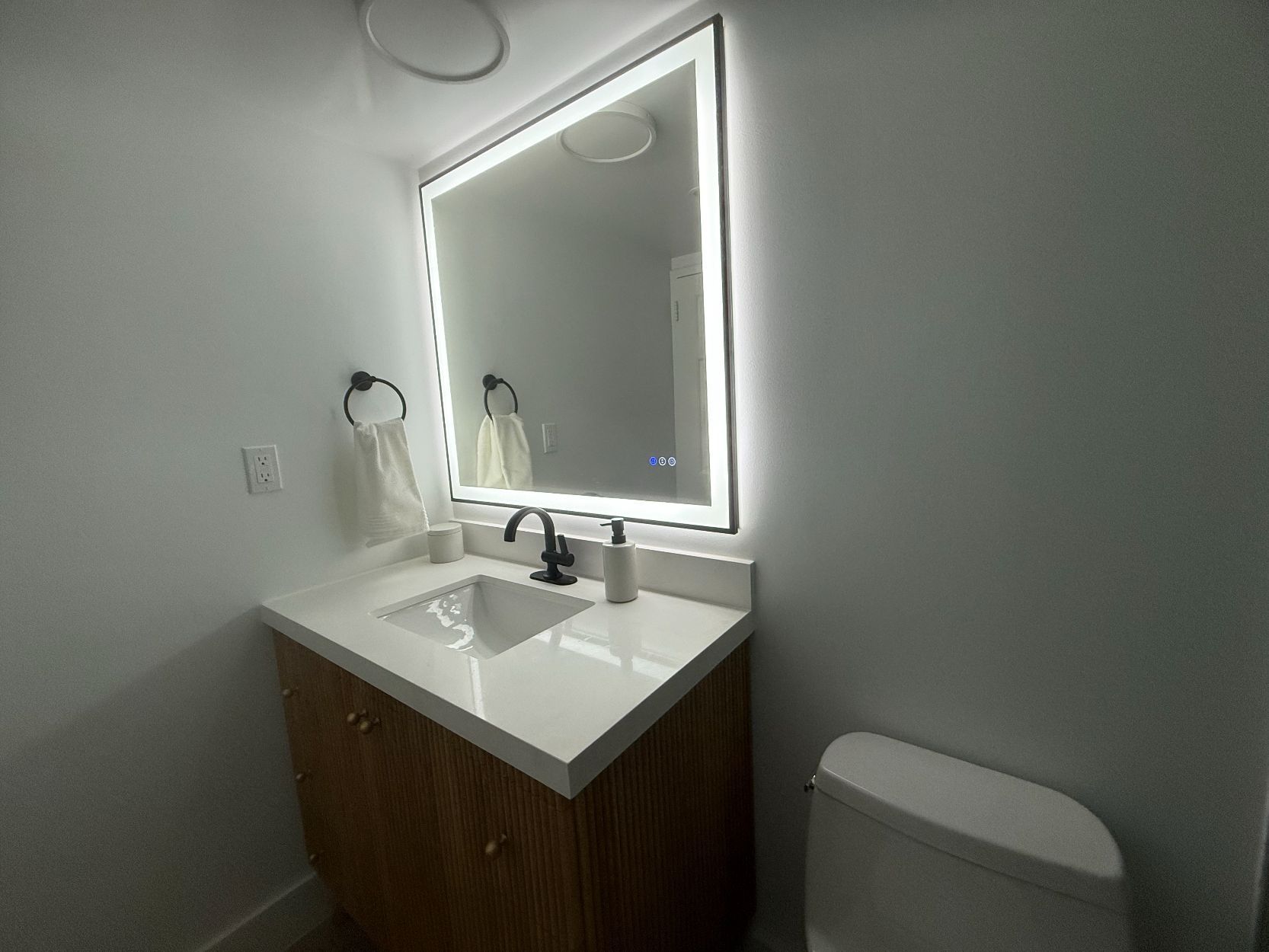 Modern bathroom vanity with lit mirror, white sink, and wood cabinet next to a toilet.