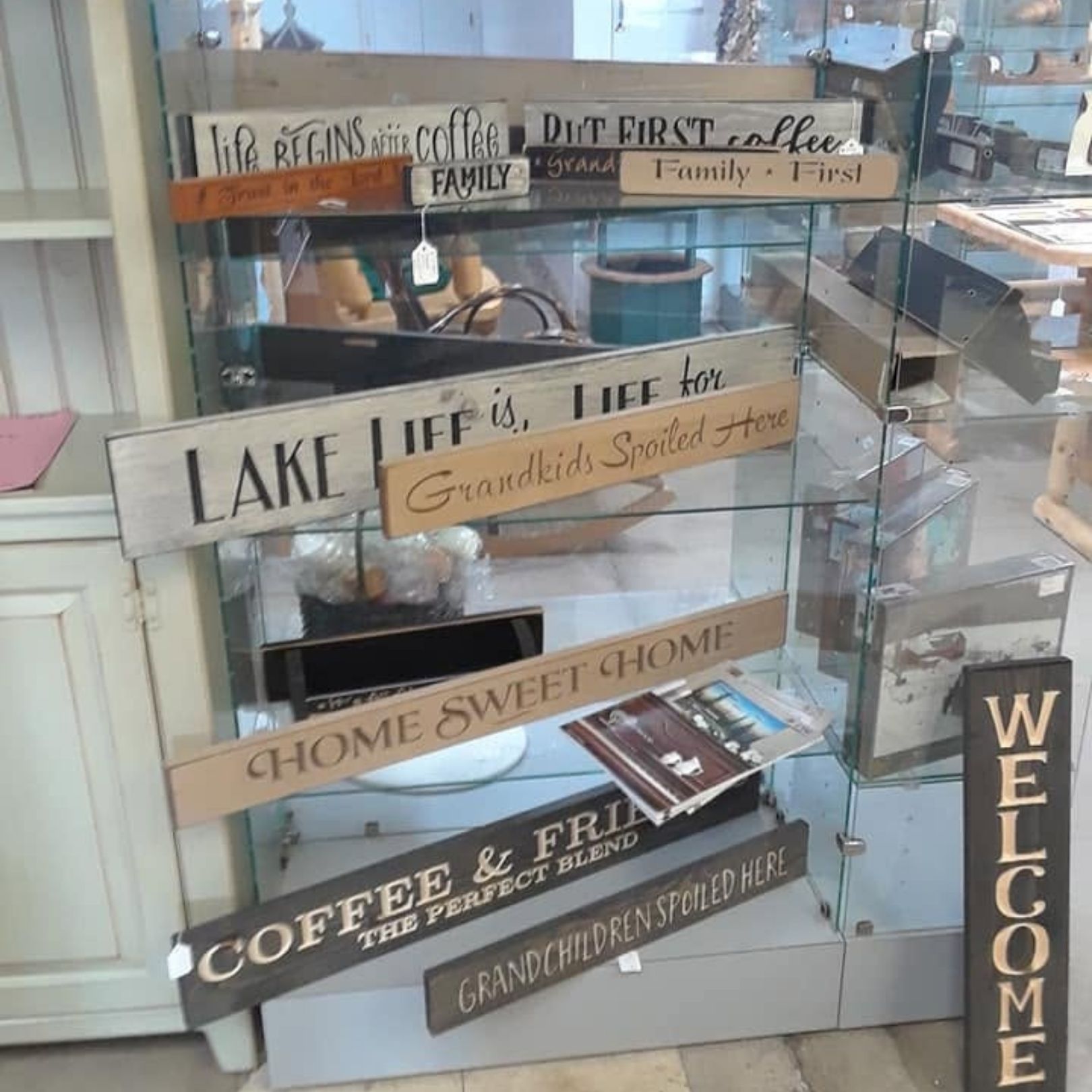 Stacked wooden home decor signs in a shop window, with phrases like “Lake Life,” “Home Sweet Home,” and “Welcome”