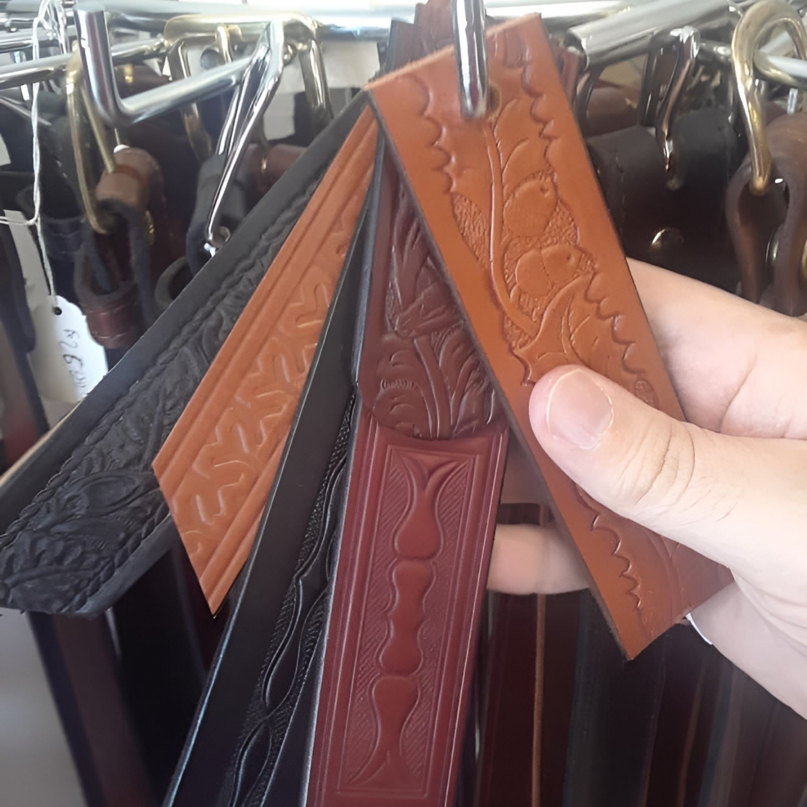 Hand holding embossed leather bag straps in black, tan, and burgundy at a clothing rack