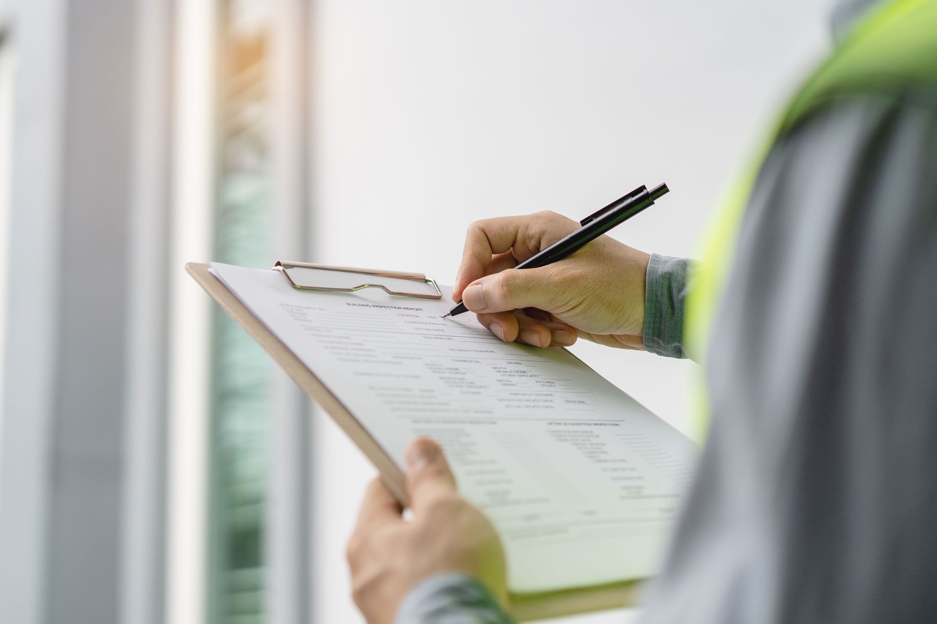 Person in a safety vest writing on a clipboard outdoors near a window.