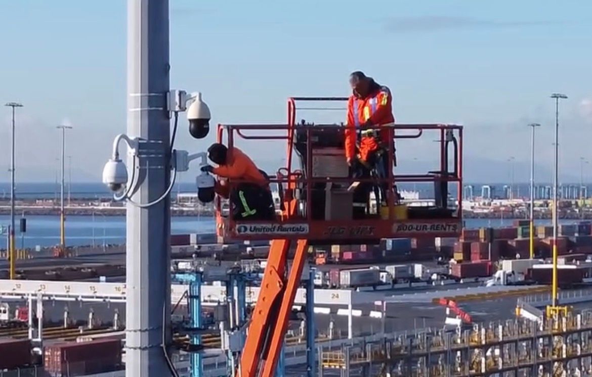 Two workers on a lift beside a tall pole, installing equipment at an industrial site near water.