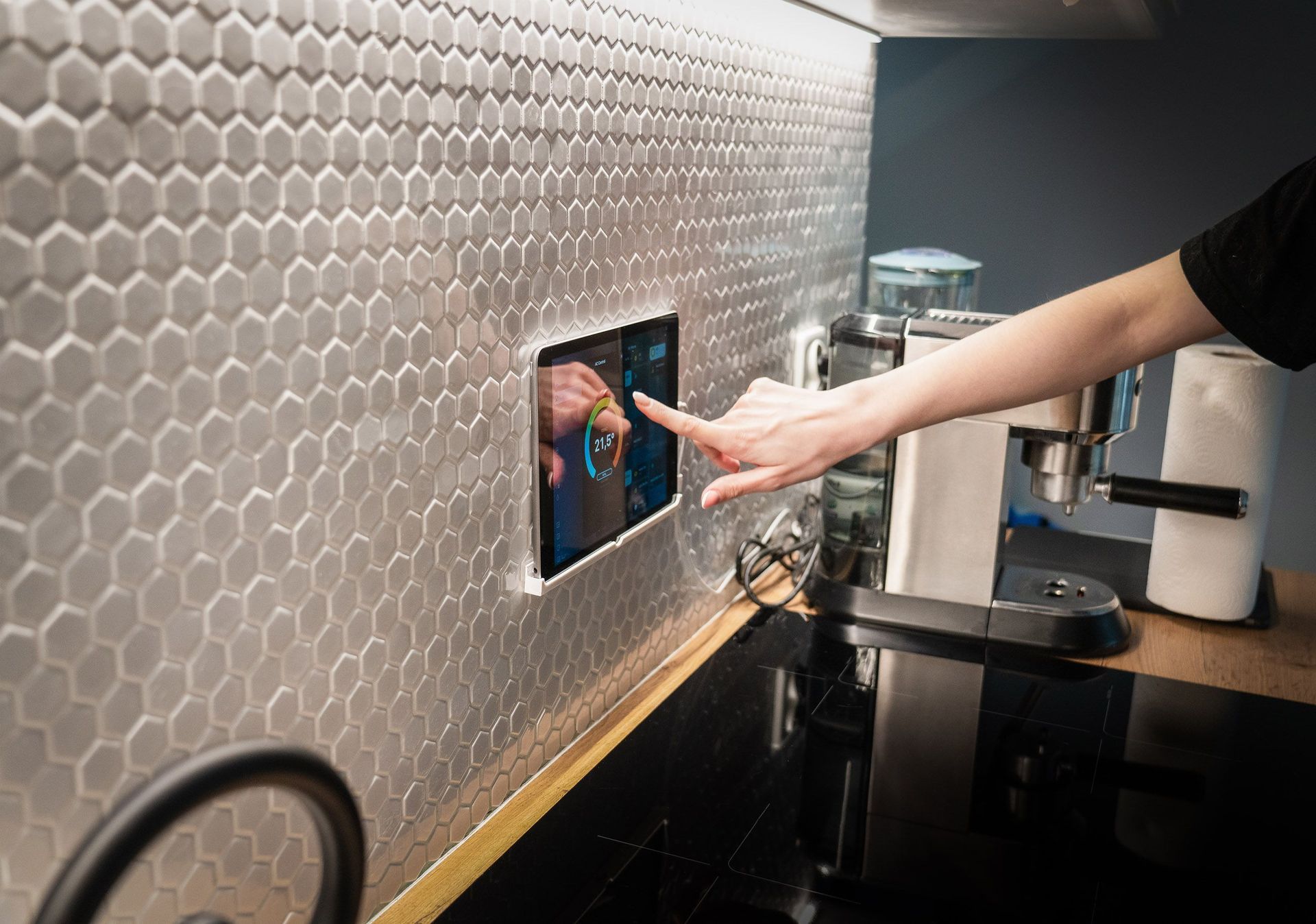 Hand pressing a wall-mounted touchscreen in a modern kitchen with hexagon tile backsplash