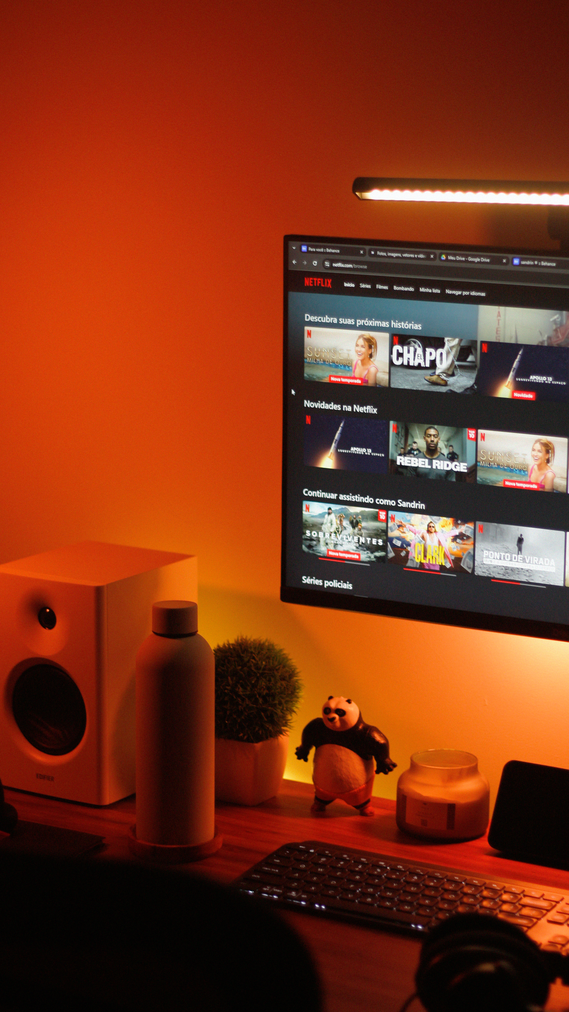 Warm orange-lit desk with monitor showing streaming app, speakers, keyboard, and small decor items.