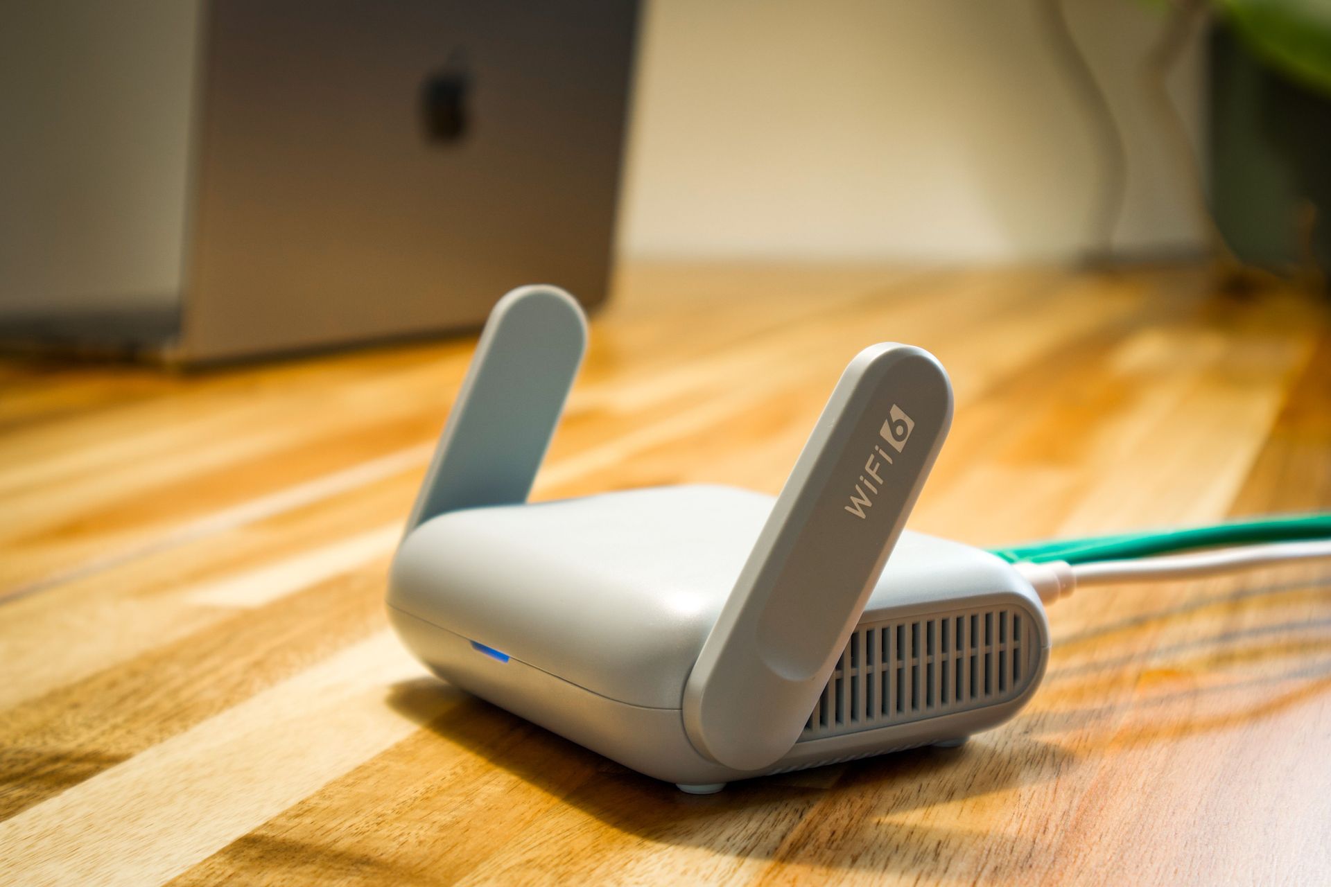 White Wi‑Fi router with two antennas on a wooden table, with a laptop blurred in the background