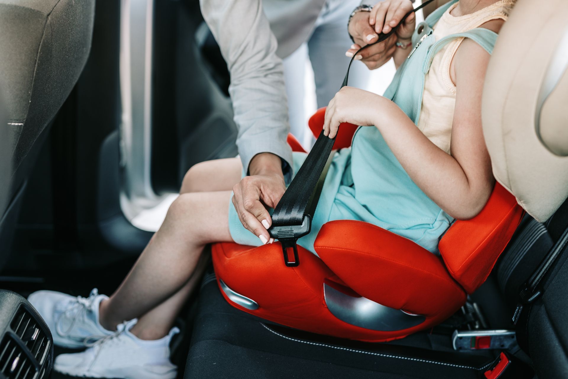 An adult helping a child buckle a seatbelt while seated in a red booster car seat inside a vehicle.