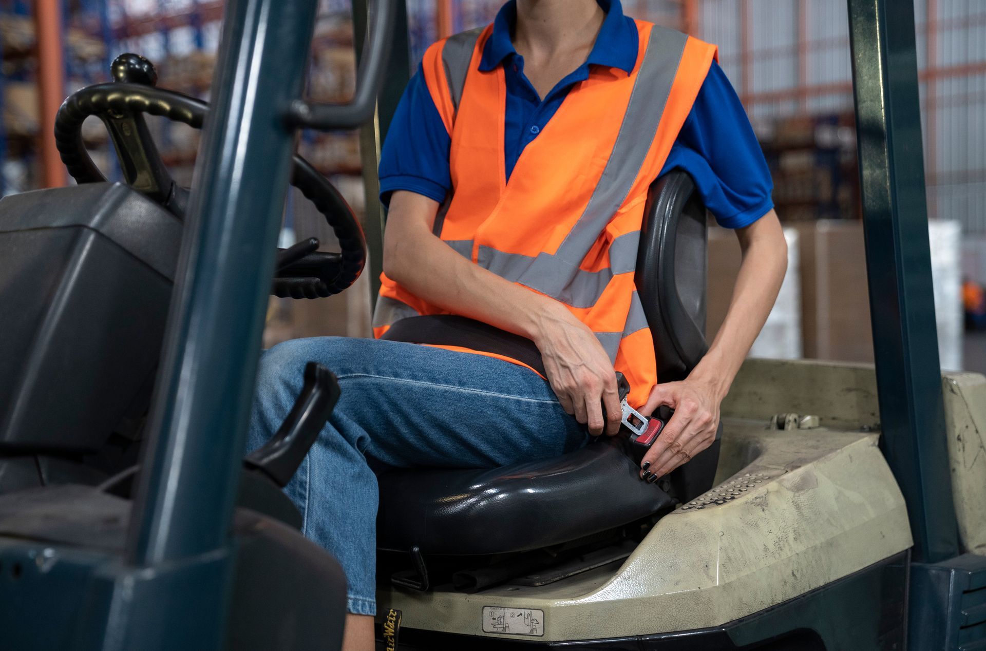 An operator in a blue shirt and high-visibility vest buckles their seatbelt while sitting in a warehouse forklift.