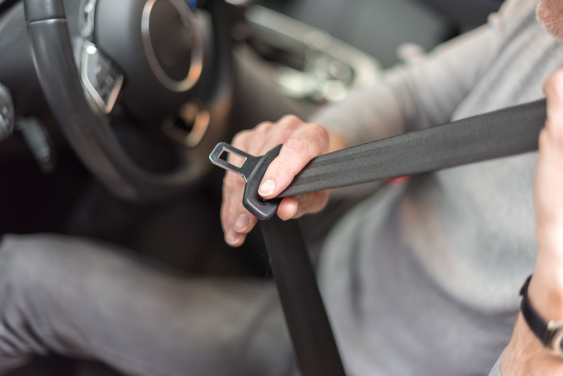 A person sits in the driver's seat of a car, holding and pulling a black seatbelt across their torso to fasten it.