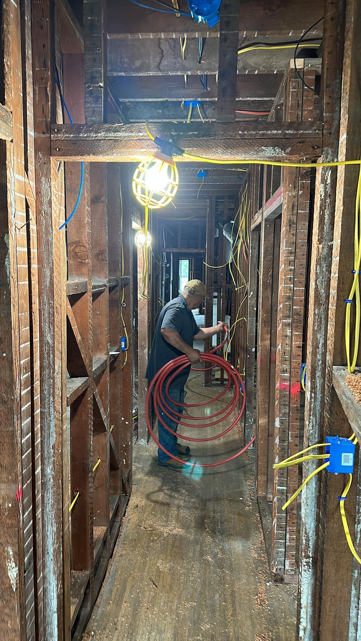 Man working on electrical wiring in a narrow hallway; exposed studs, red tubing, blue boxes, yellow wires, bright light.