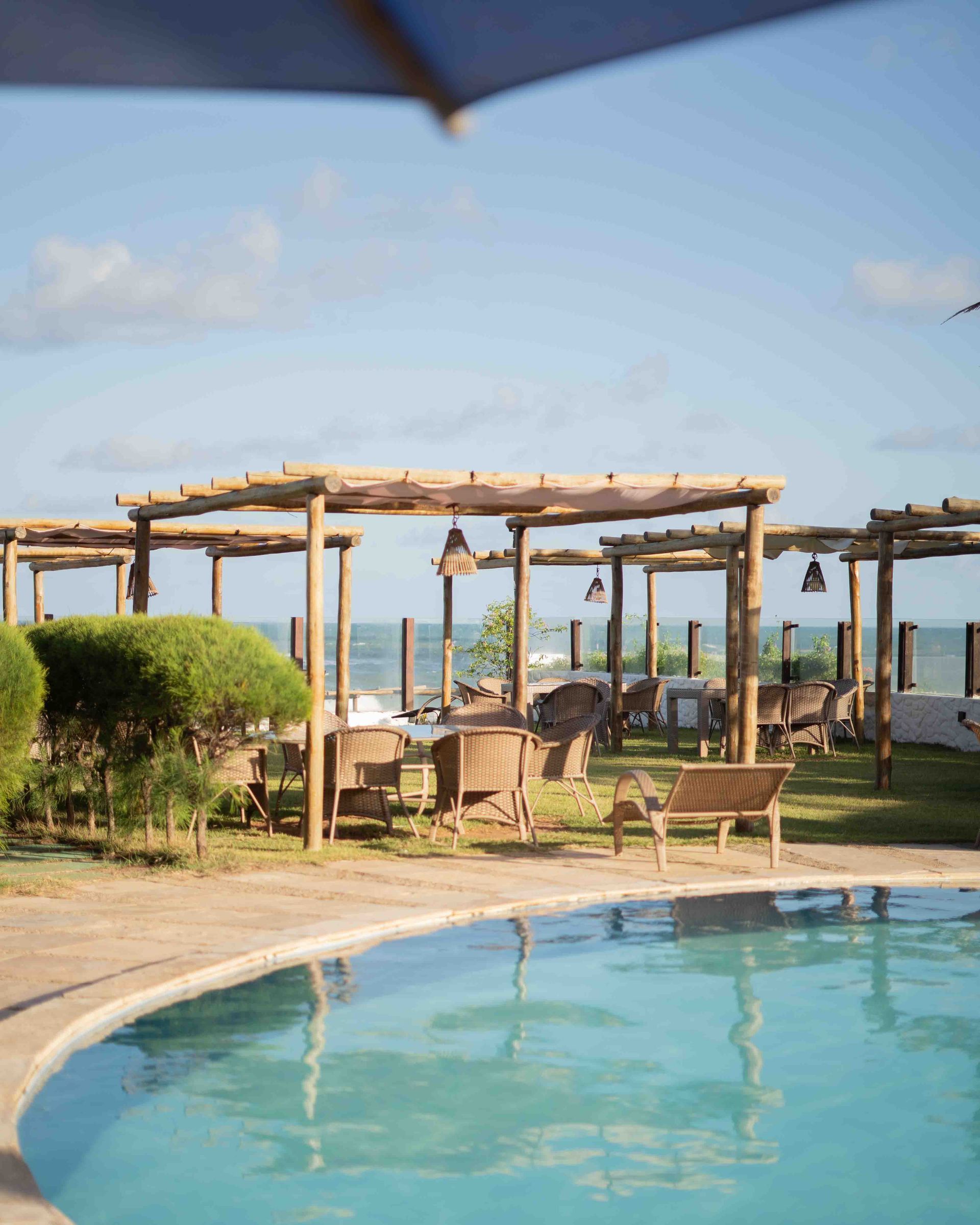 Piscina com vista para a área de refeições ao ar livre perto do oceano, sob um céu azul.