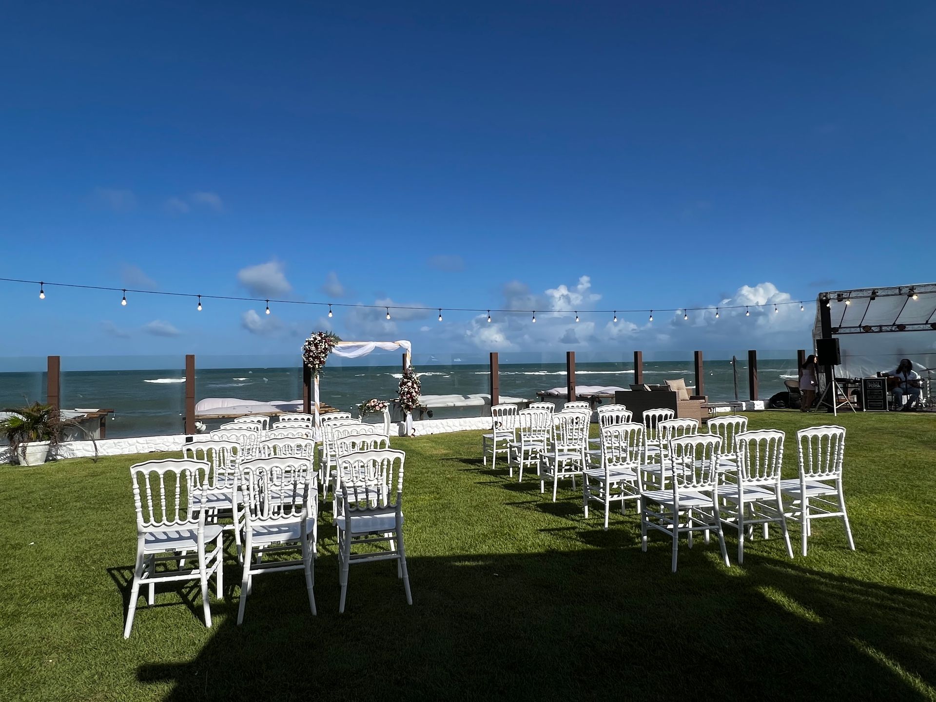 Cerimônia de casamento montada em gramado verde com vista para o oceano. Cadeiras brancas de frente para um arco.