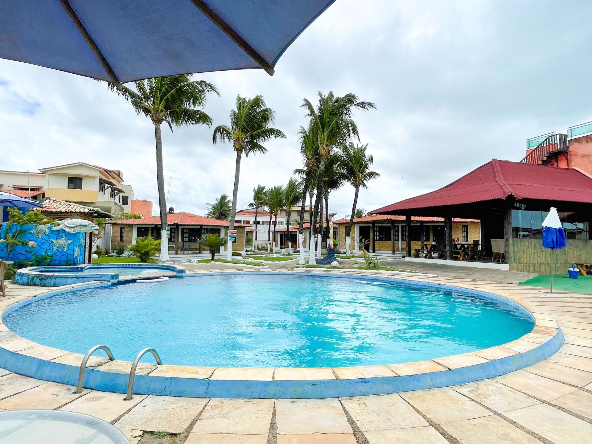 Piscina em um resort com palmeiras, prédios e céu nublado.