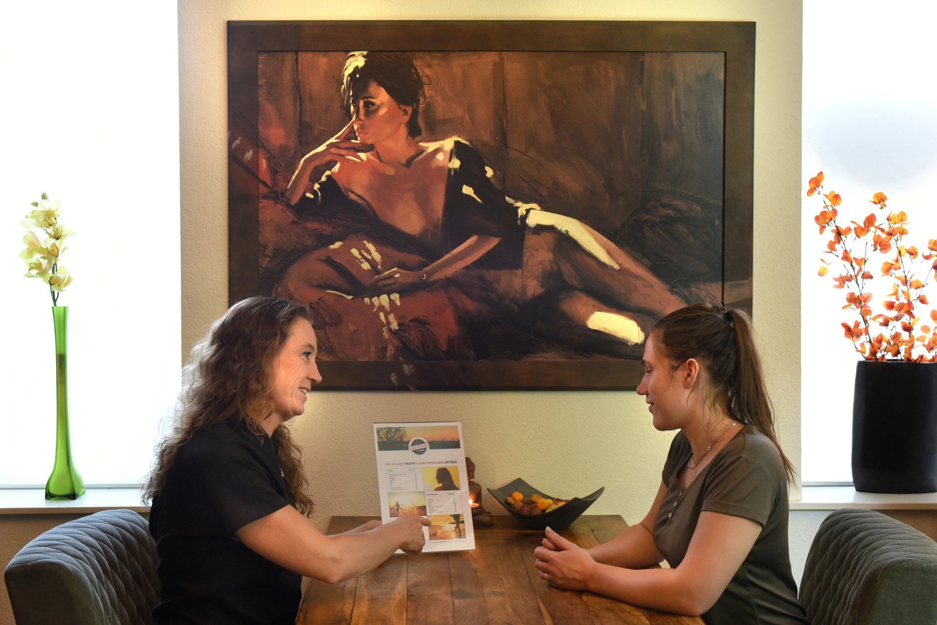 Twee mensen zitten aan een houten tafel in een kantoorachtige ruimte en bespreken een brochure voor een schilderij van een liggende vrouw.