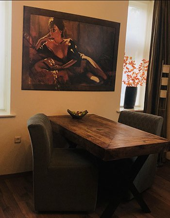 Een eethoek met een rustieke houten tafel, twee grijze gestoffeerde stoelen en een groot schilderij van een figuur aan de muur.