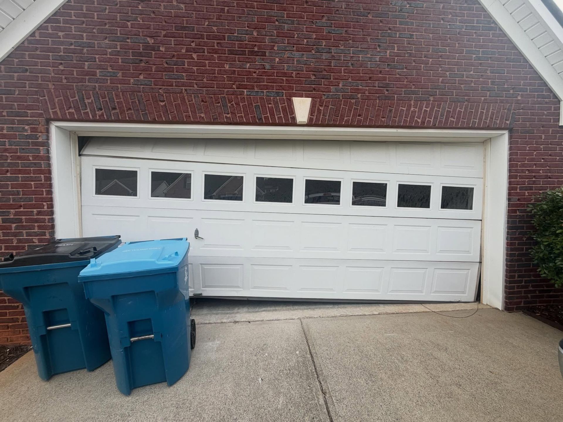 Two blue trash bins in front of a white garage door on a brick house driveway