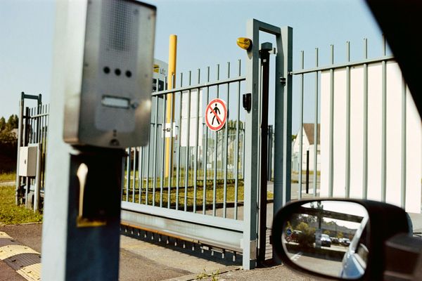 Industrial gate with metal fence and warning sign, seen from a car window.