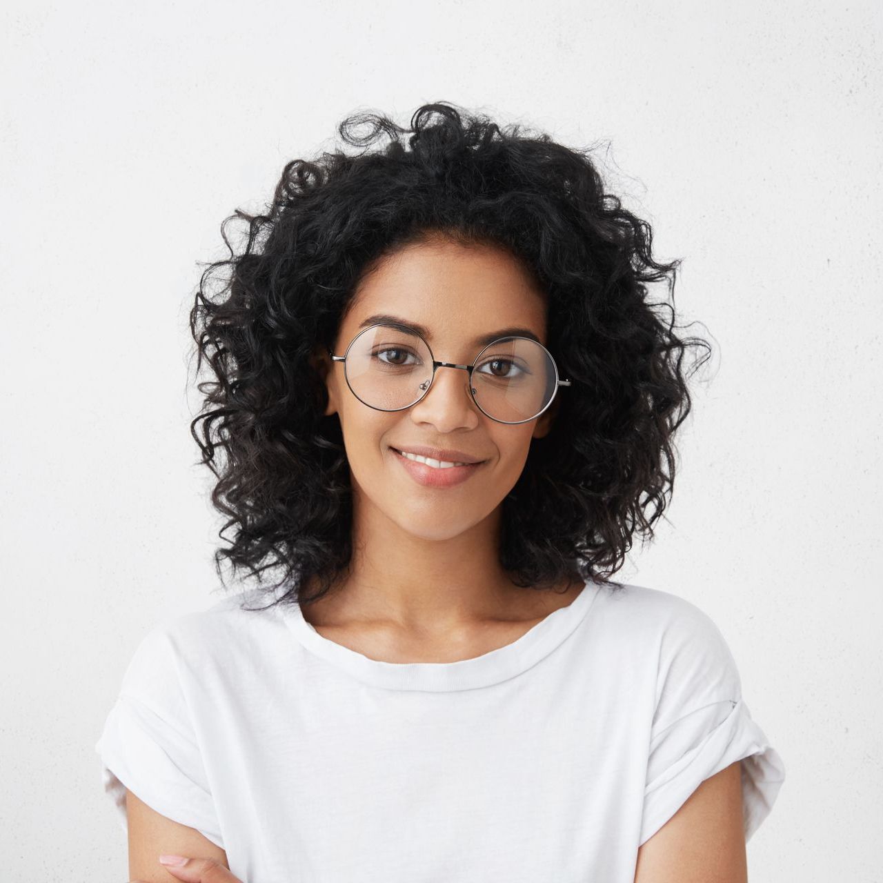 Woman with dark curly hair and glasses smiling, wearing a white t-shirt.