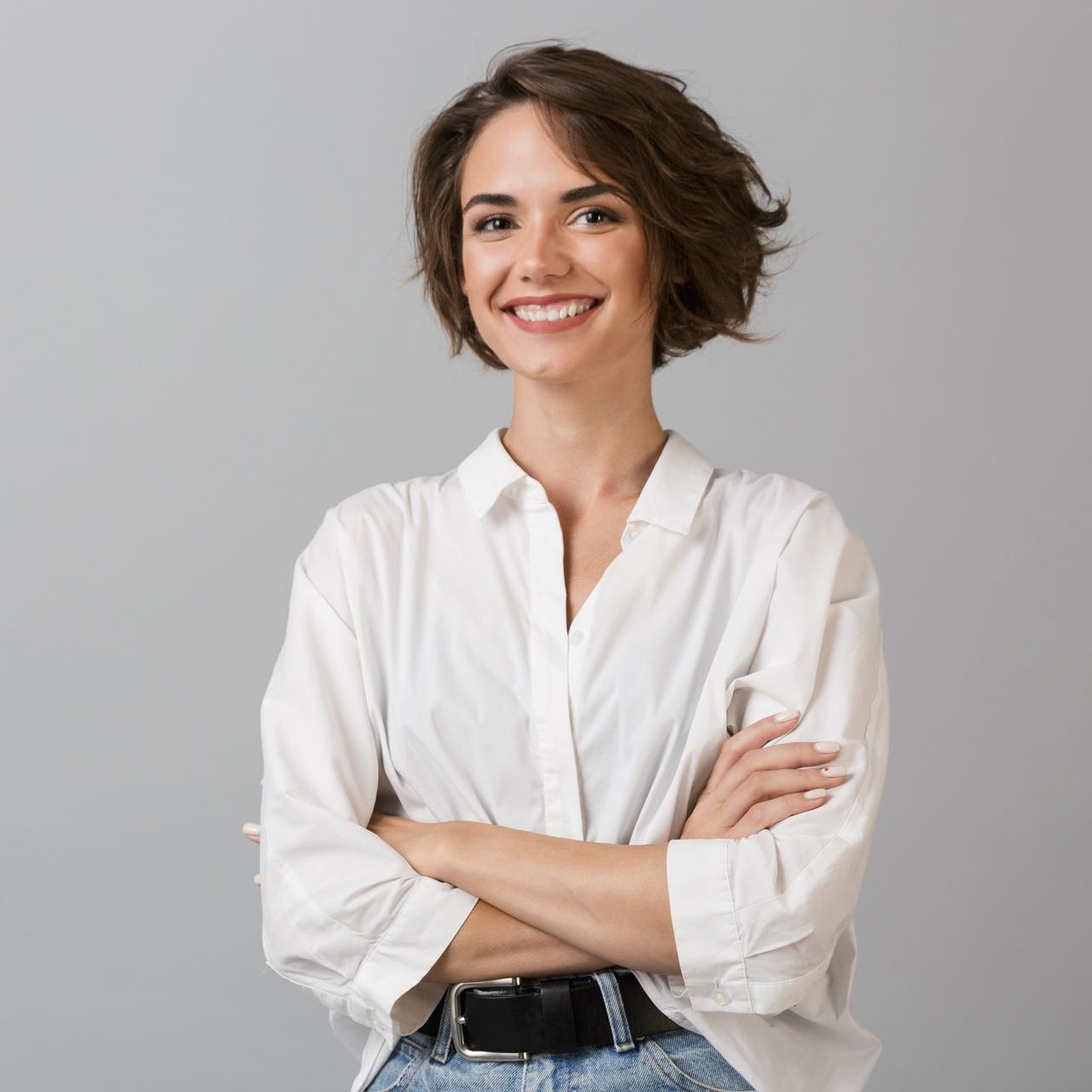 Woman with short brown hair, smiling, arms crossed, wearing a white shirt and jeans against a gray backdrop.