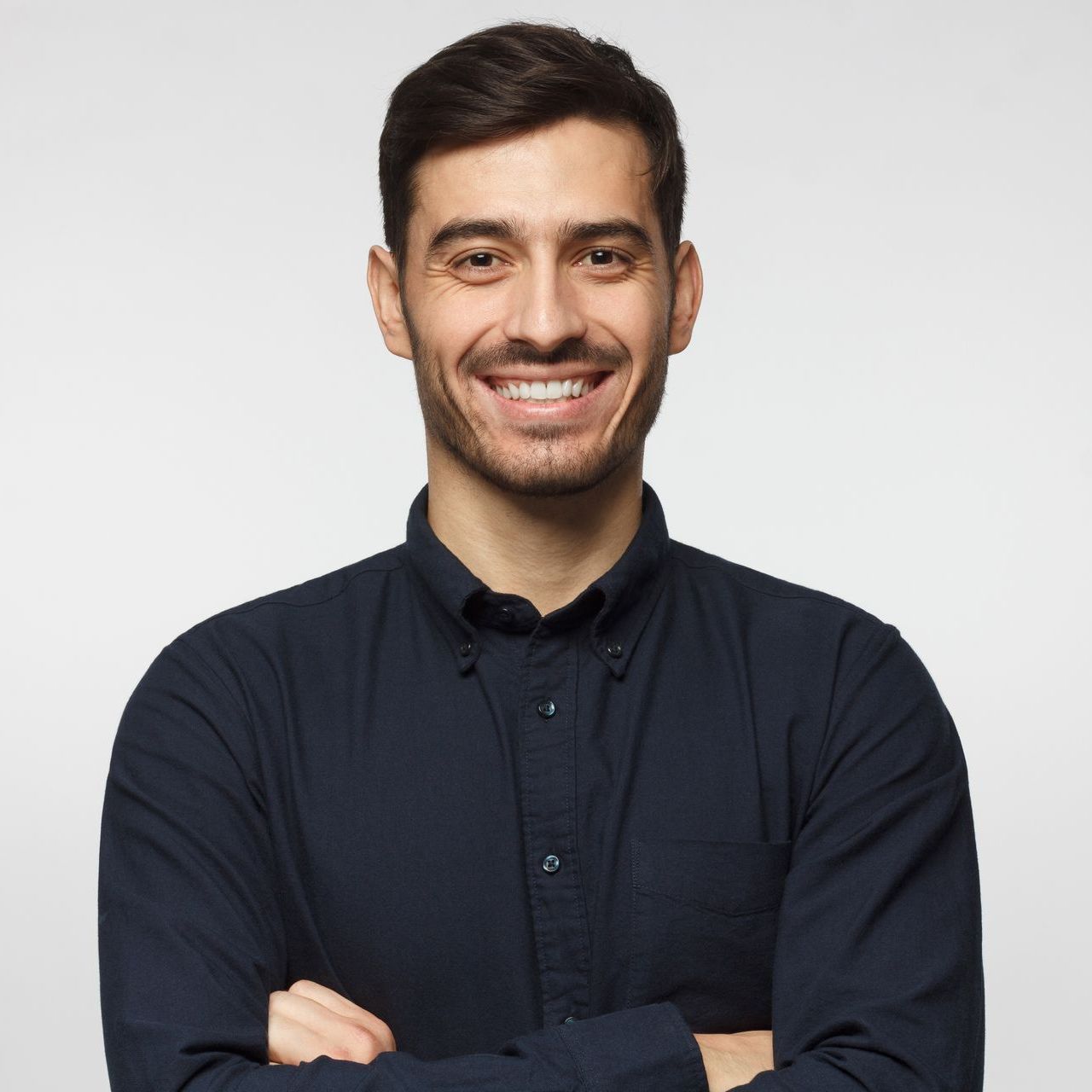 Man with dark hair and a beard smiles with arms crossed, wearing a dark blue button-up shirt against a gray background.