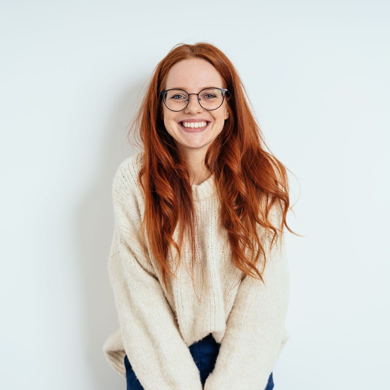 Woman with red hair and glasses smiles, wearing a cream sweater against a white wall.