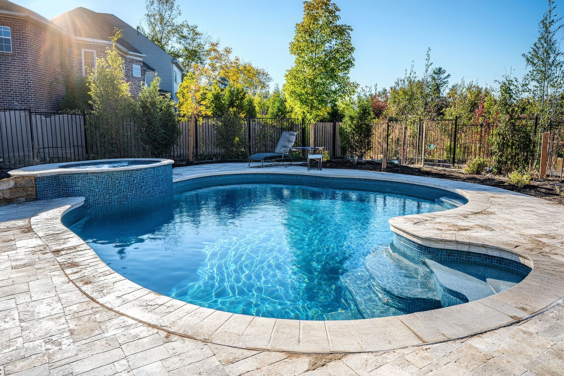 Backyard pool with blue water and stone surround; hot tub in the corner and trees in the background.