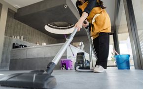 Person vacuuming a modern kitchen with a silver vacuum, wearing a mustard jacket and black pants.