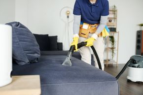 Person vacuuming a gray sofa in a bright living room with a handheld upholstery cleaner
