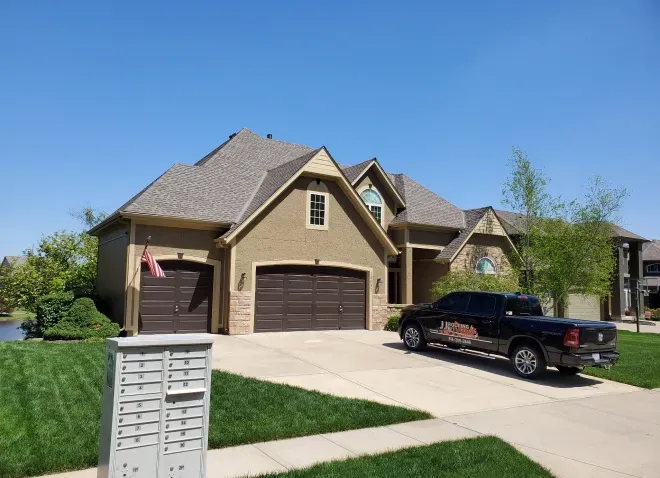 A tan two-story house with a brown garage and roof, featuring a black pickup truck parked in the driveway on a sunny day.