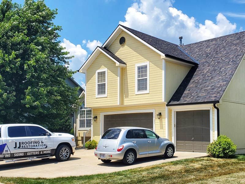 A pale yellow, two-story house with two garage doors, a silver hatchback, and a white work truck in the driveway.