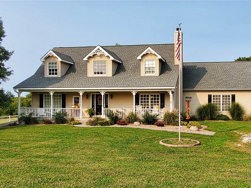 A beige, two-story house with a front porch, three dormer windows, a gray shingled roof, and an American flag on a pole.