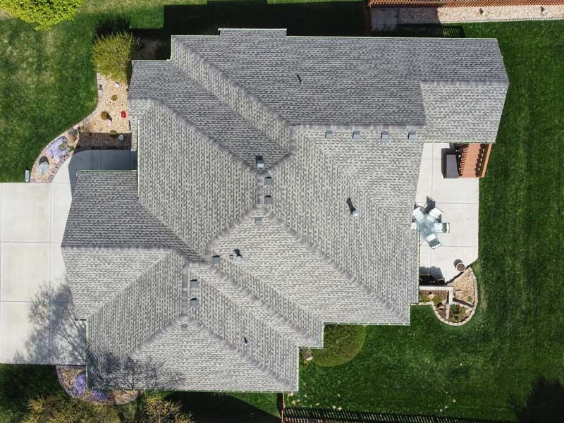 An aerial top-down view of a complex, grey-shingled roof on a house with a surrounding lawn and concrete patio.