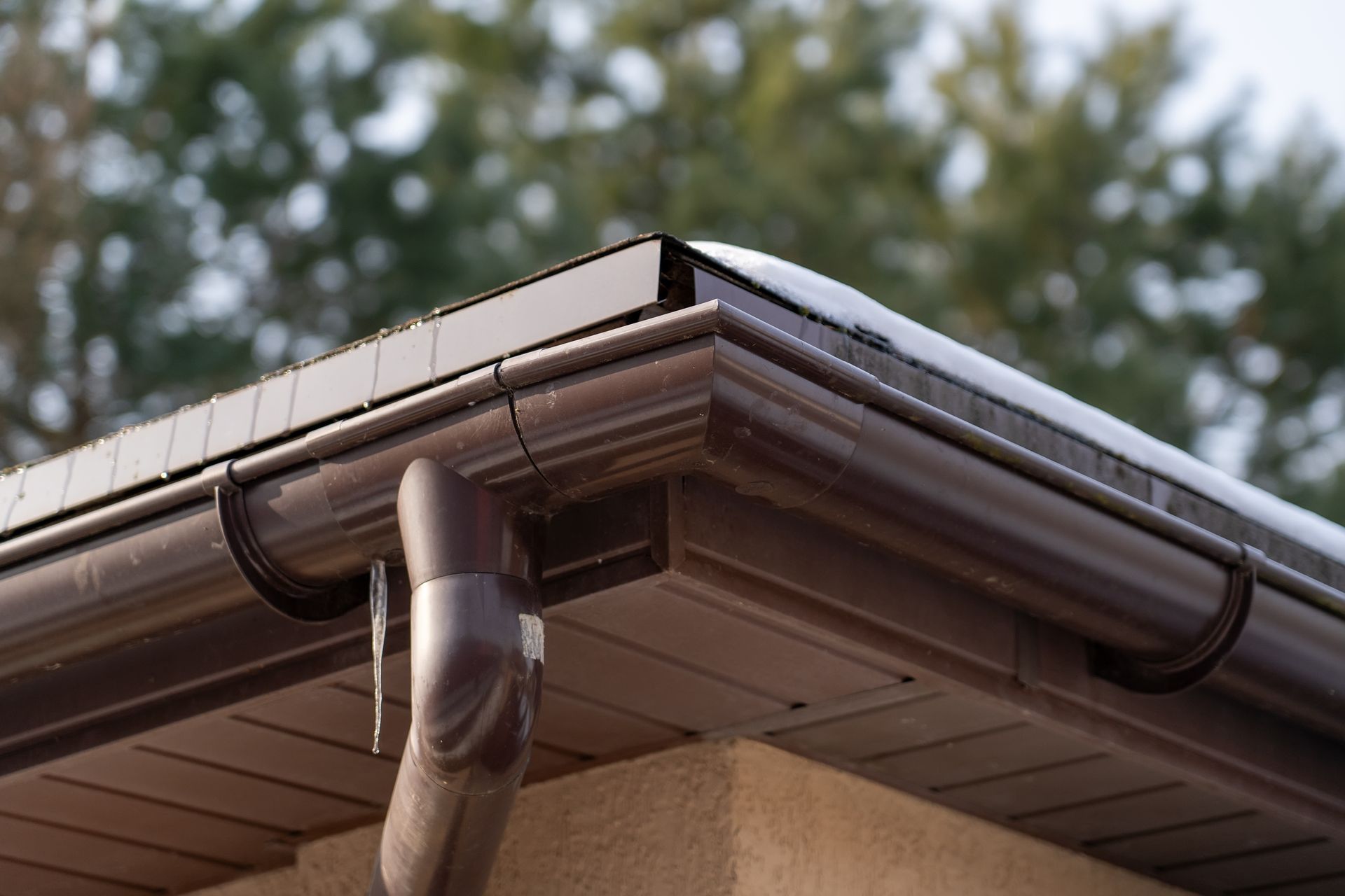 A corner of a roofline with brown gutters and downspout, featuring an icicle hanging near a patch of snow.