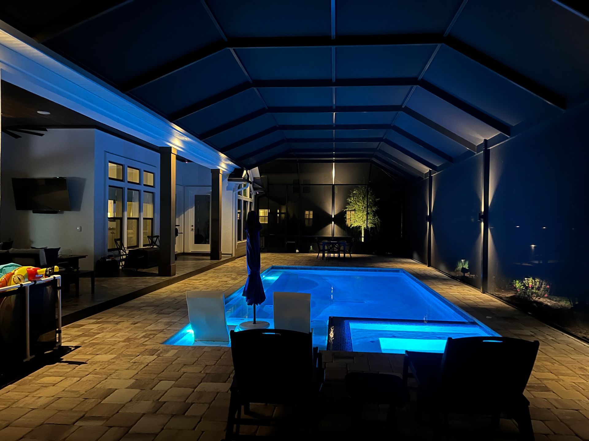 Night view of a pool area with blue illuminated pool, seating, and overhead canopy. Brick patio, trees, and house are also visible.