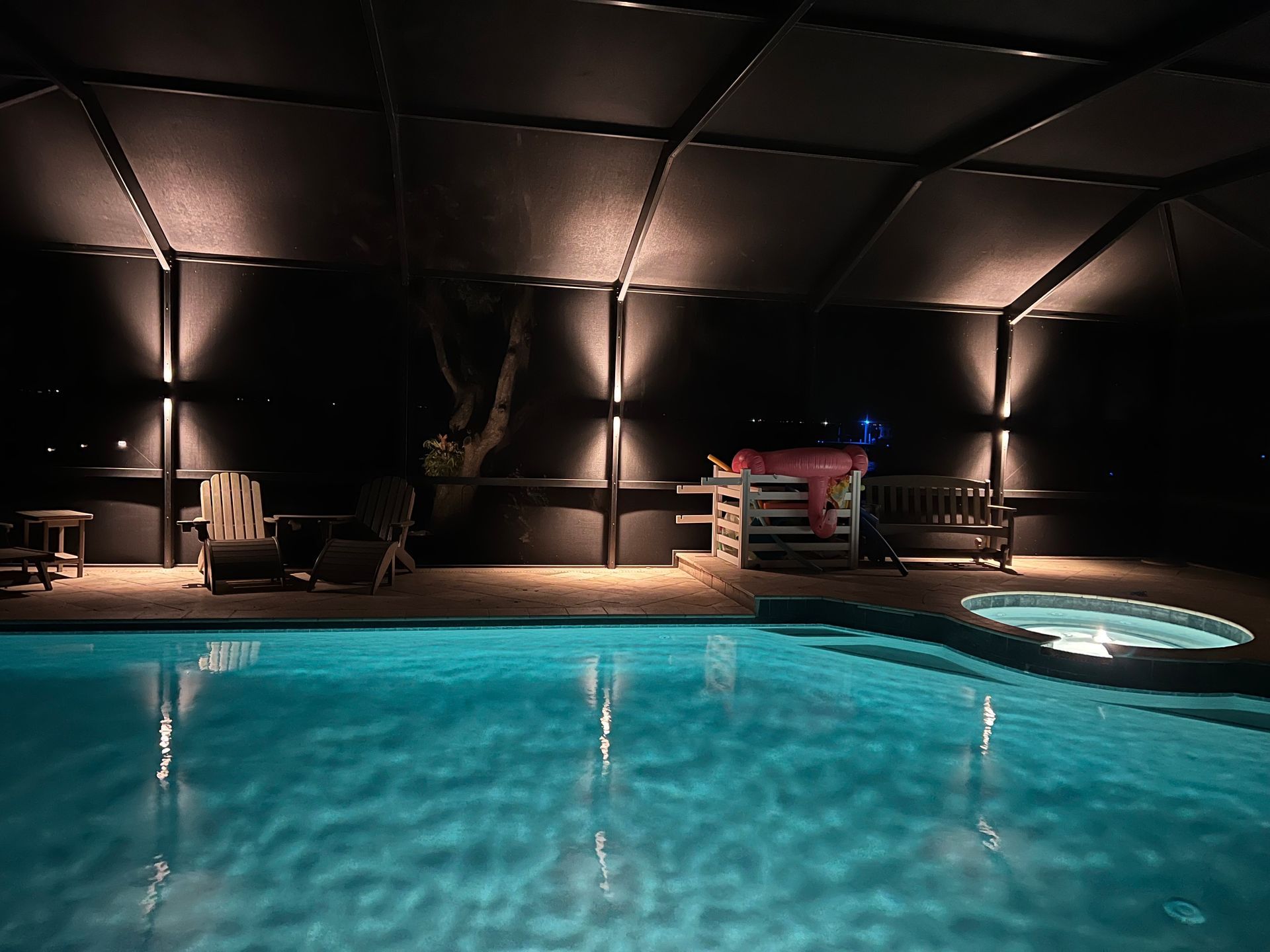 Poolside patio at night, illuminated by spotlights. Pool reflects the lights, with furniture visible.
