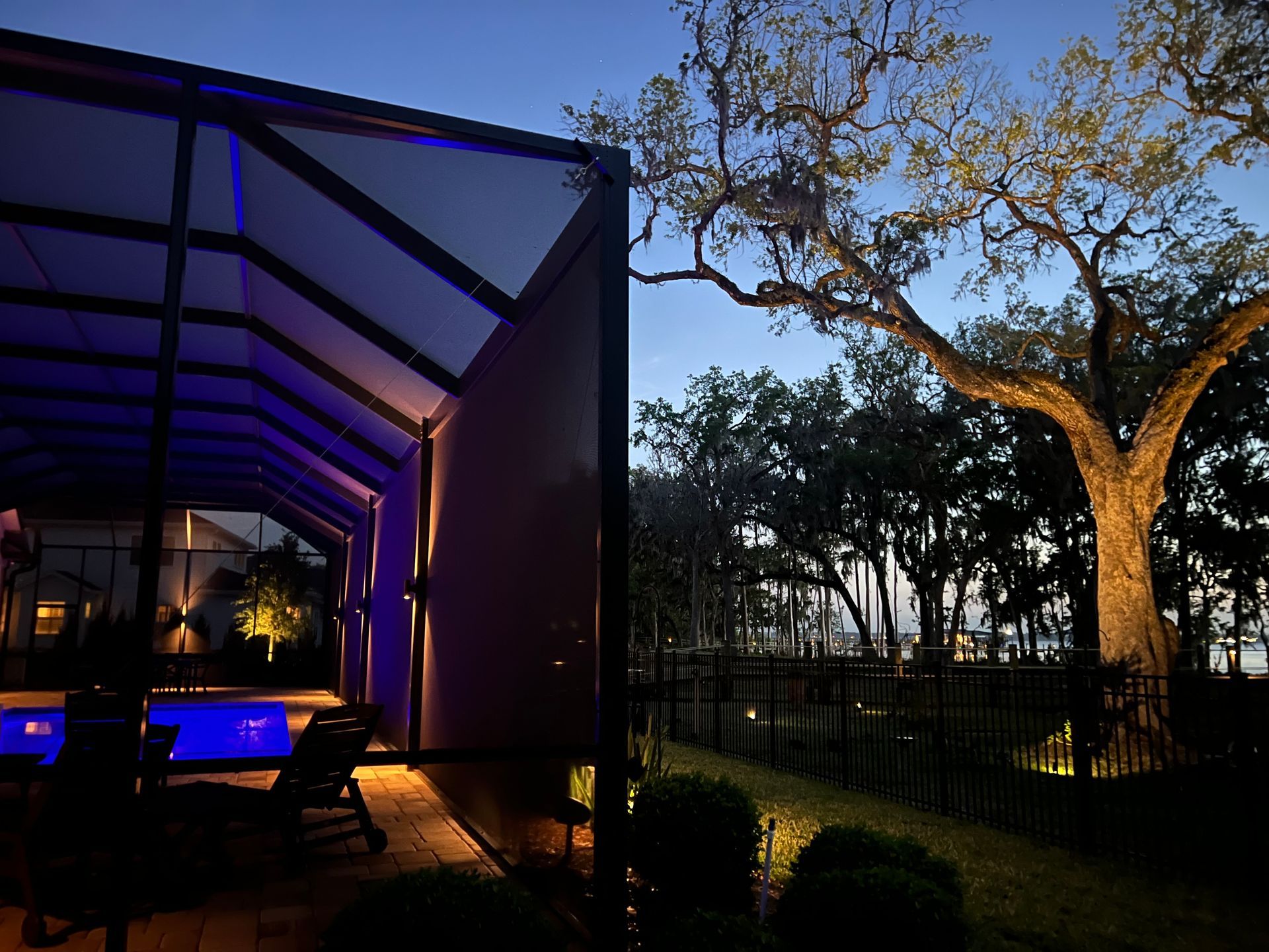 Outdoor pool area at dusk; modern architectural structure with blue lighting. Large tree in background.