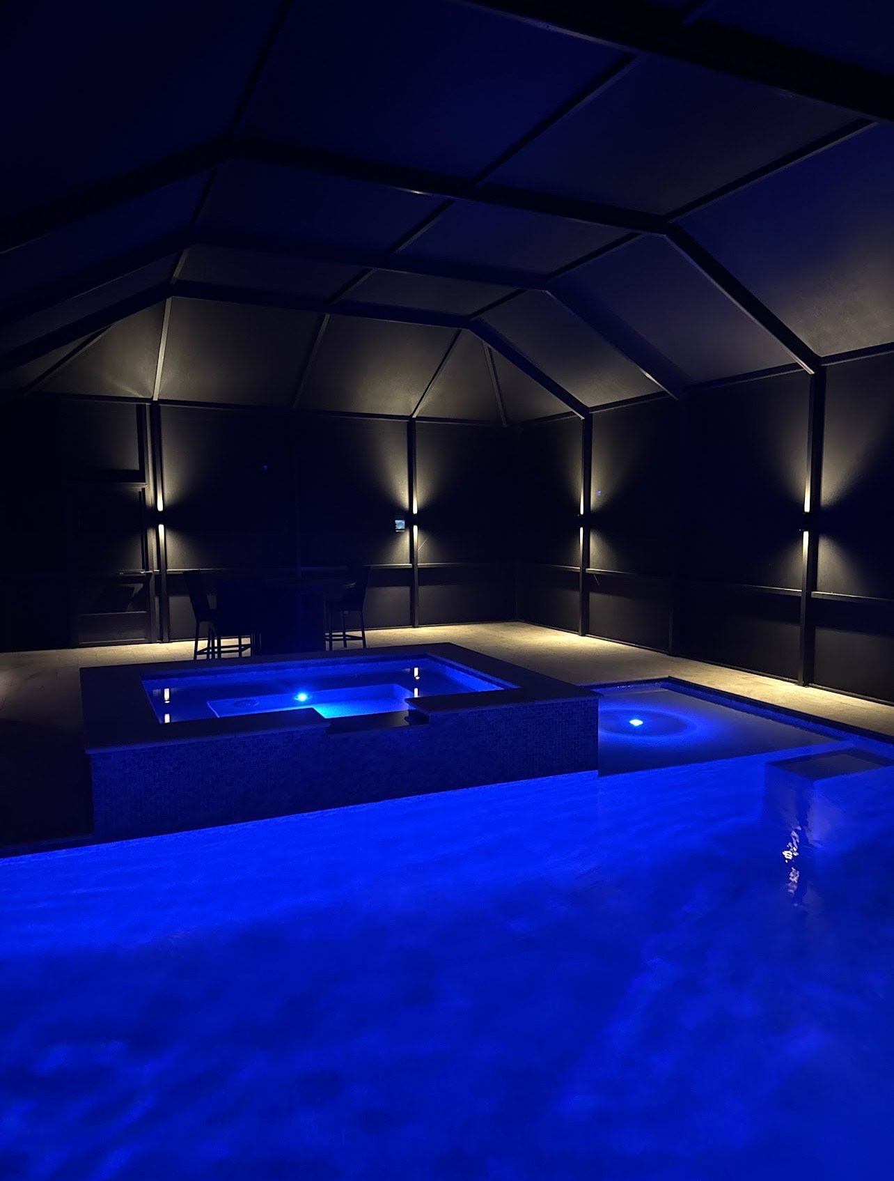 Indoor pool and jacuzzi illuminated with blue light, under a dark roof, lit by wall sconces.