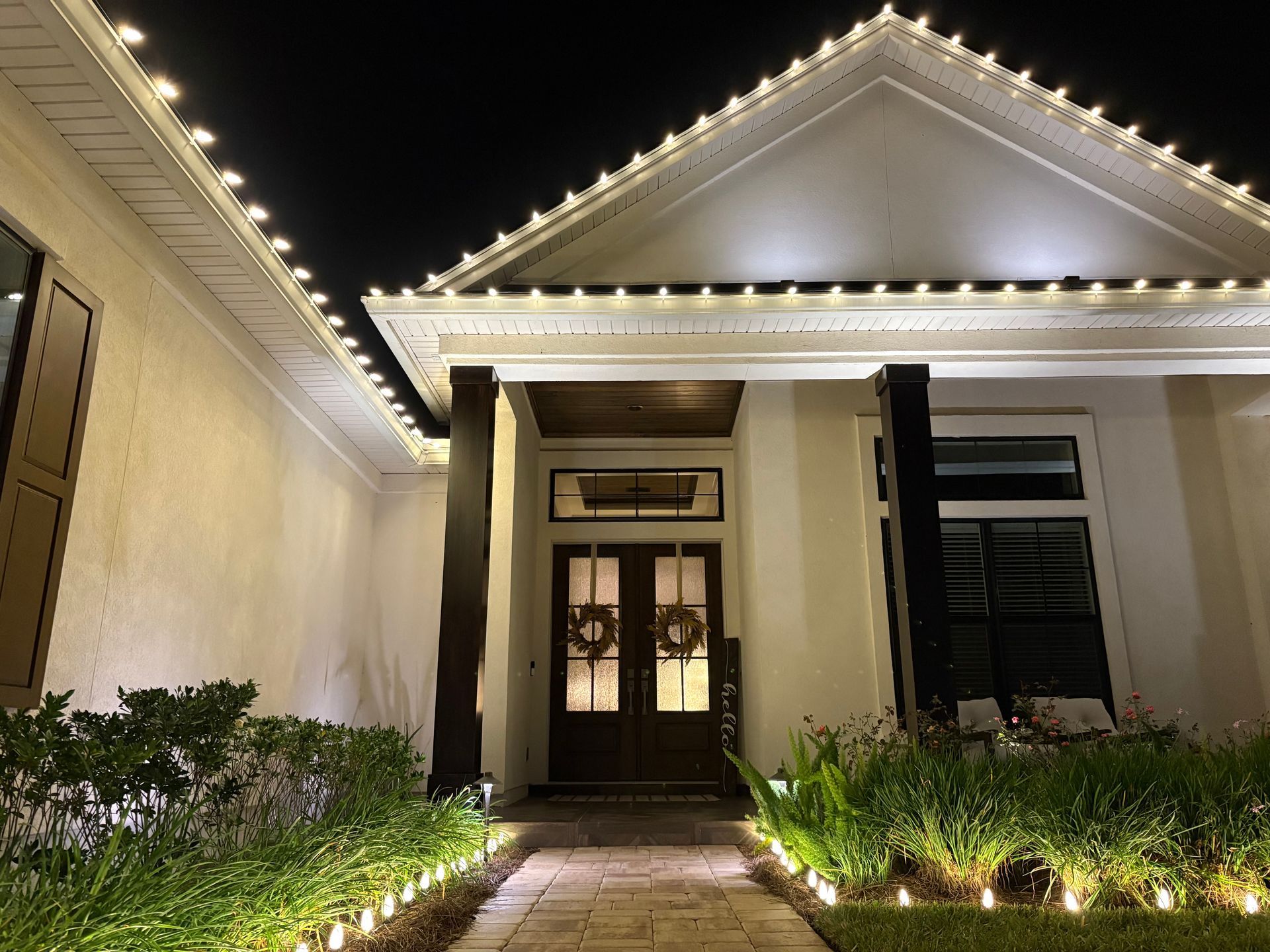 House with white walls and Christmas lights; brick pathway, bushes, and dark front door.