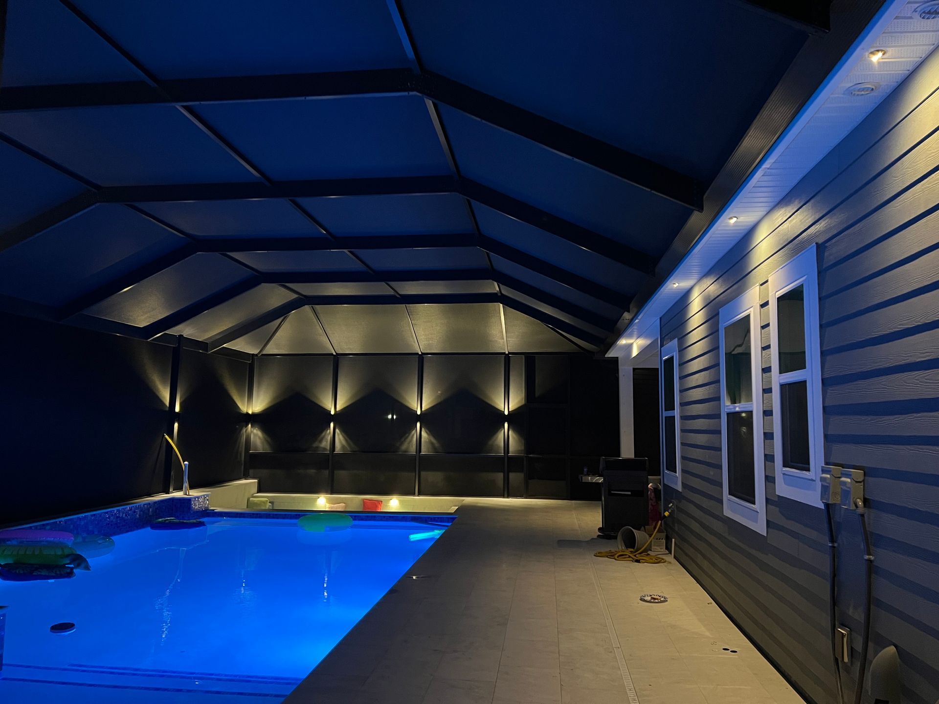 Pool area at night with blue lighting. Covered structure with recessed lights. Side of a house with windows.