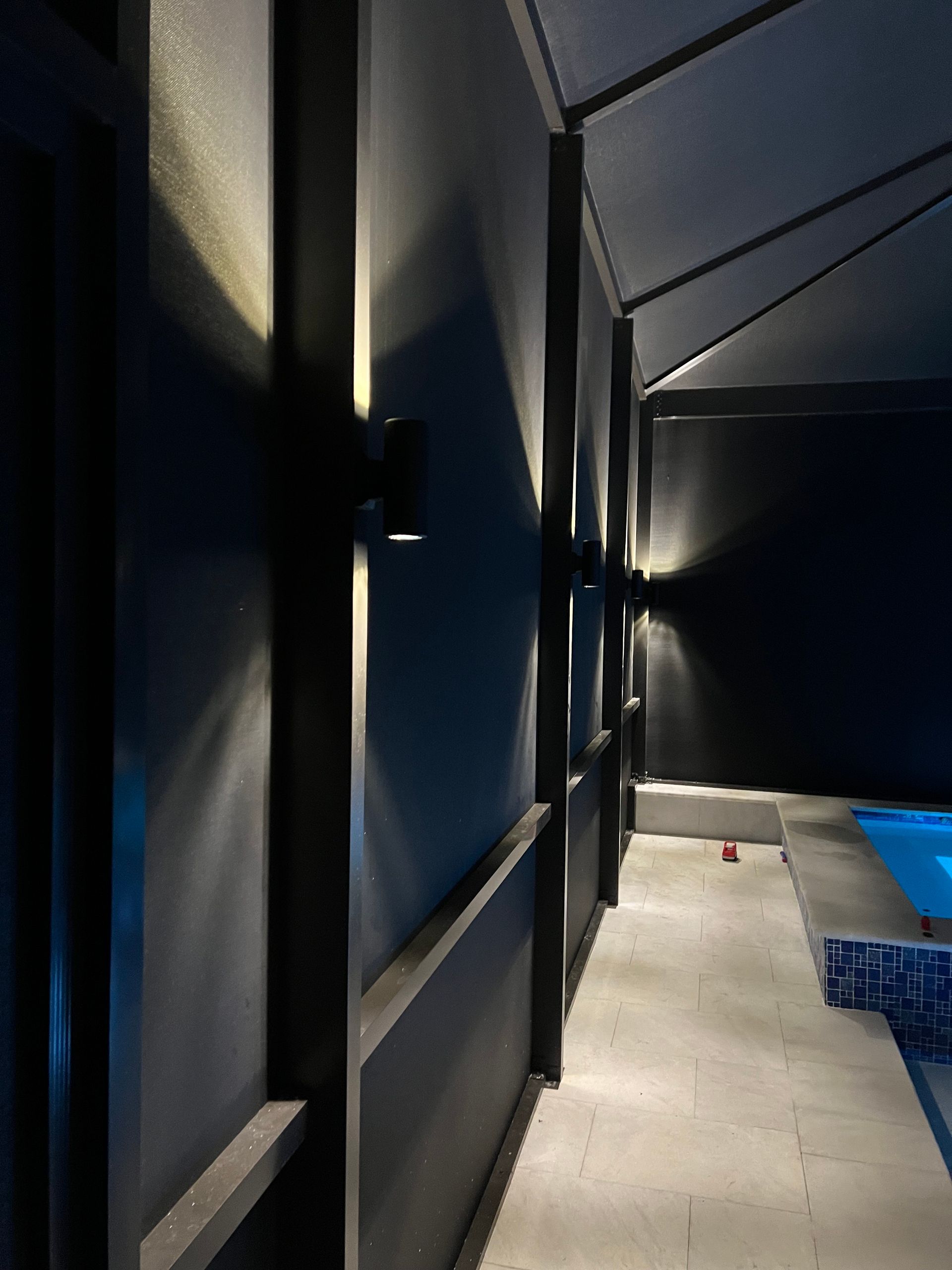 Black outdoor wall with modern sconce lights casting beams on the dark surface, alongside a pool area.