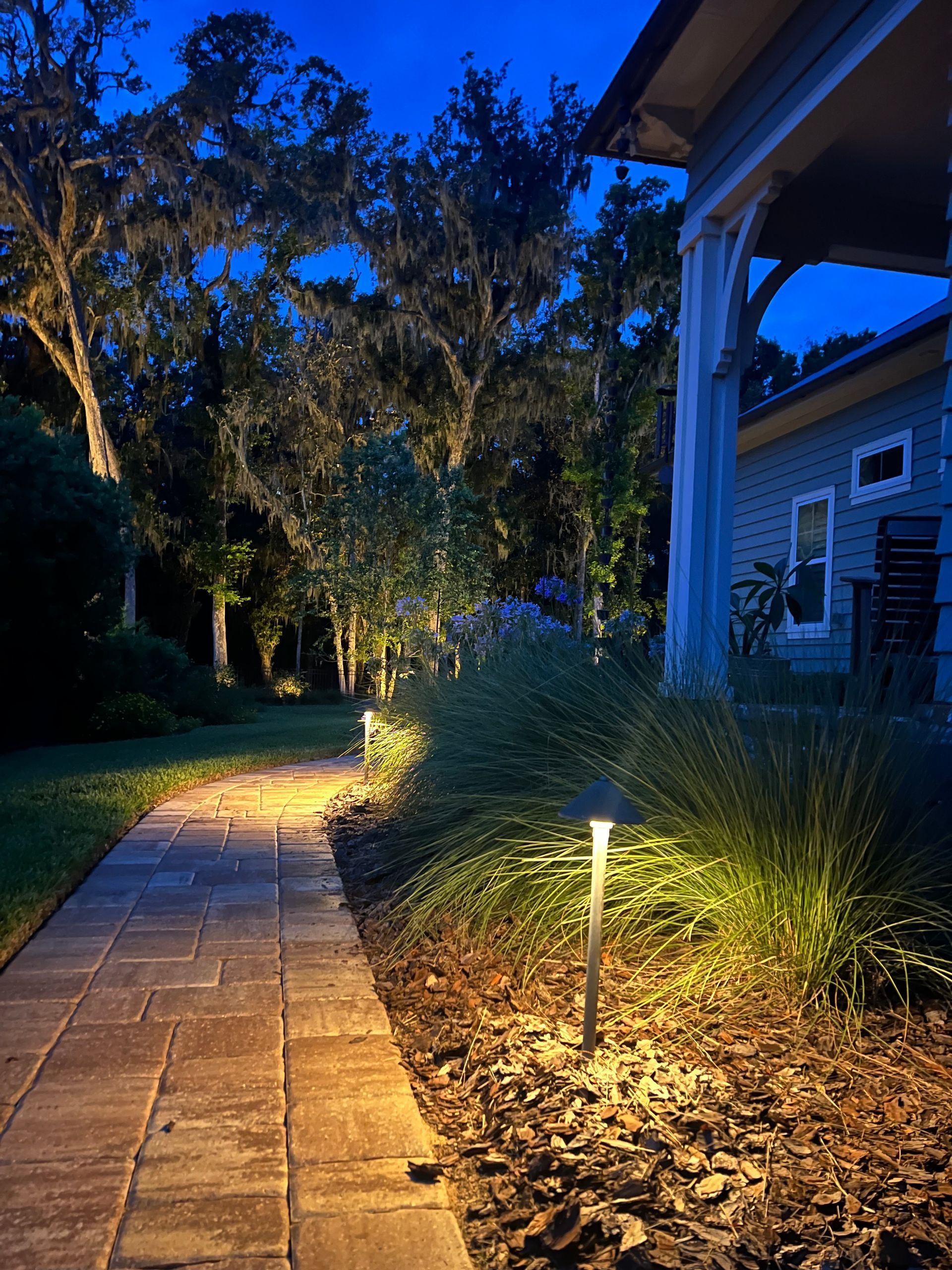 Brick pathway lit by outdoor lights leads to a house at dusk, with trees and bushes.
