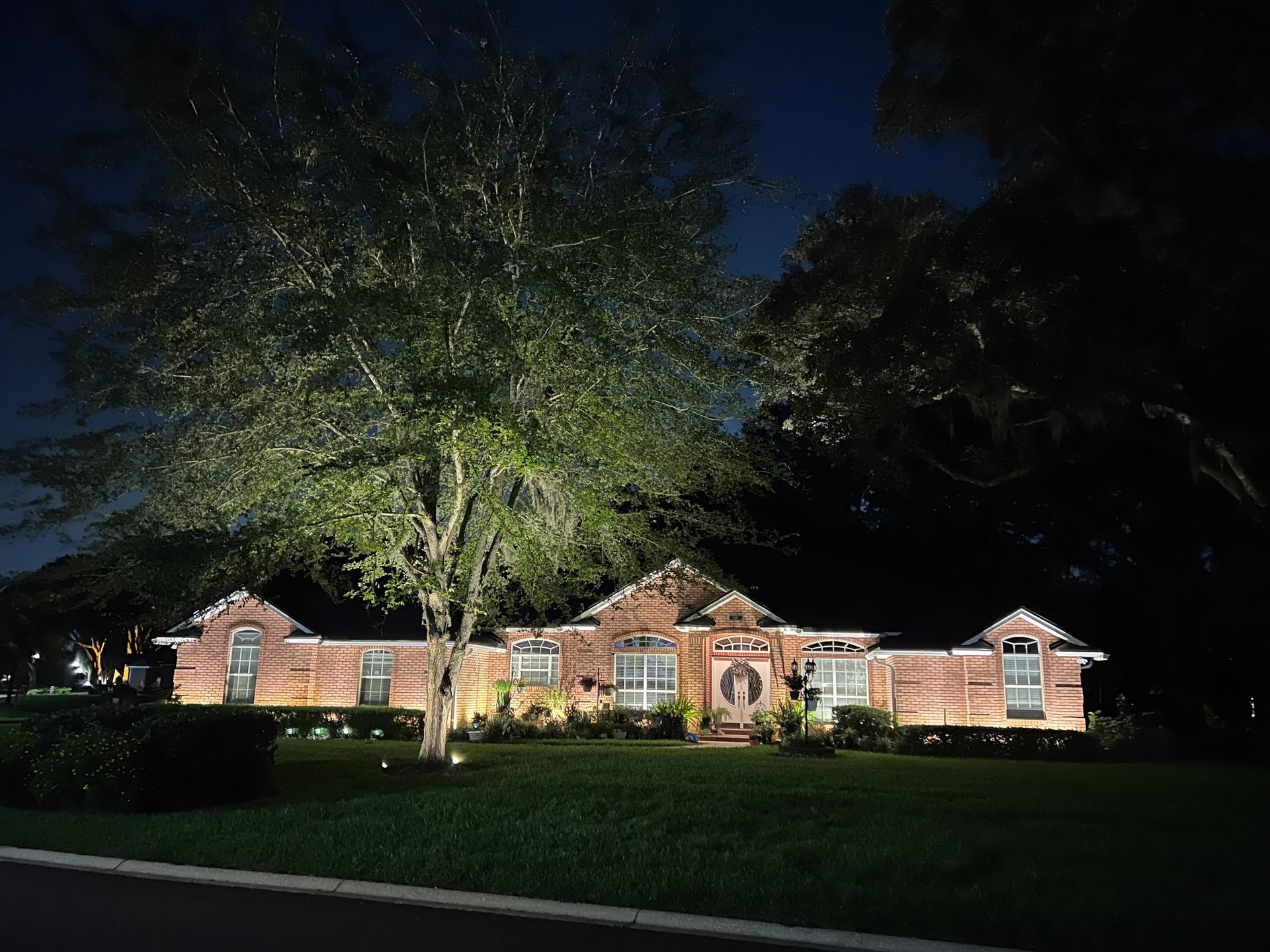 Brick house at night, illuminated by outdoor lighting, with large tree in the foreground.