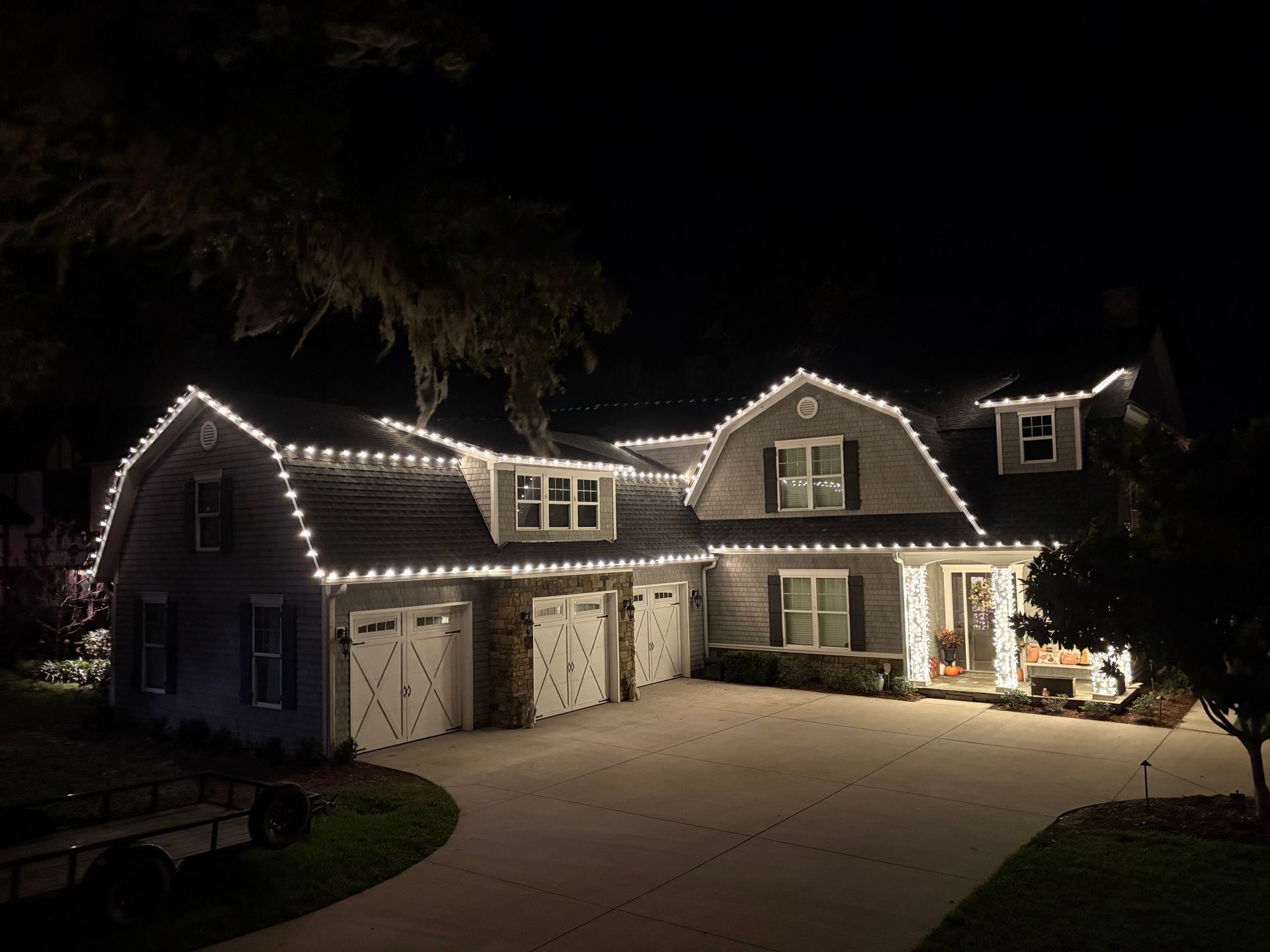 House at night with white Christmas lights outlining the roof, driveway lit, and three-car garage.