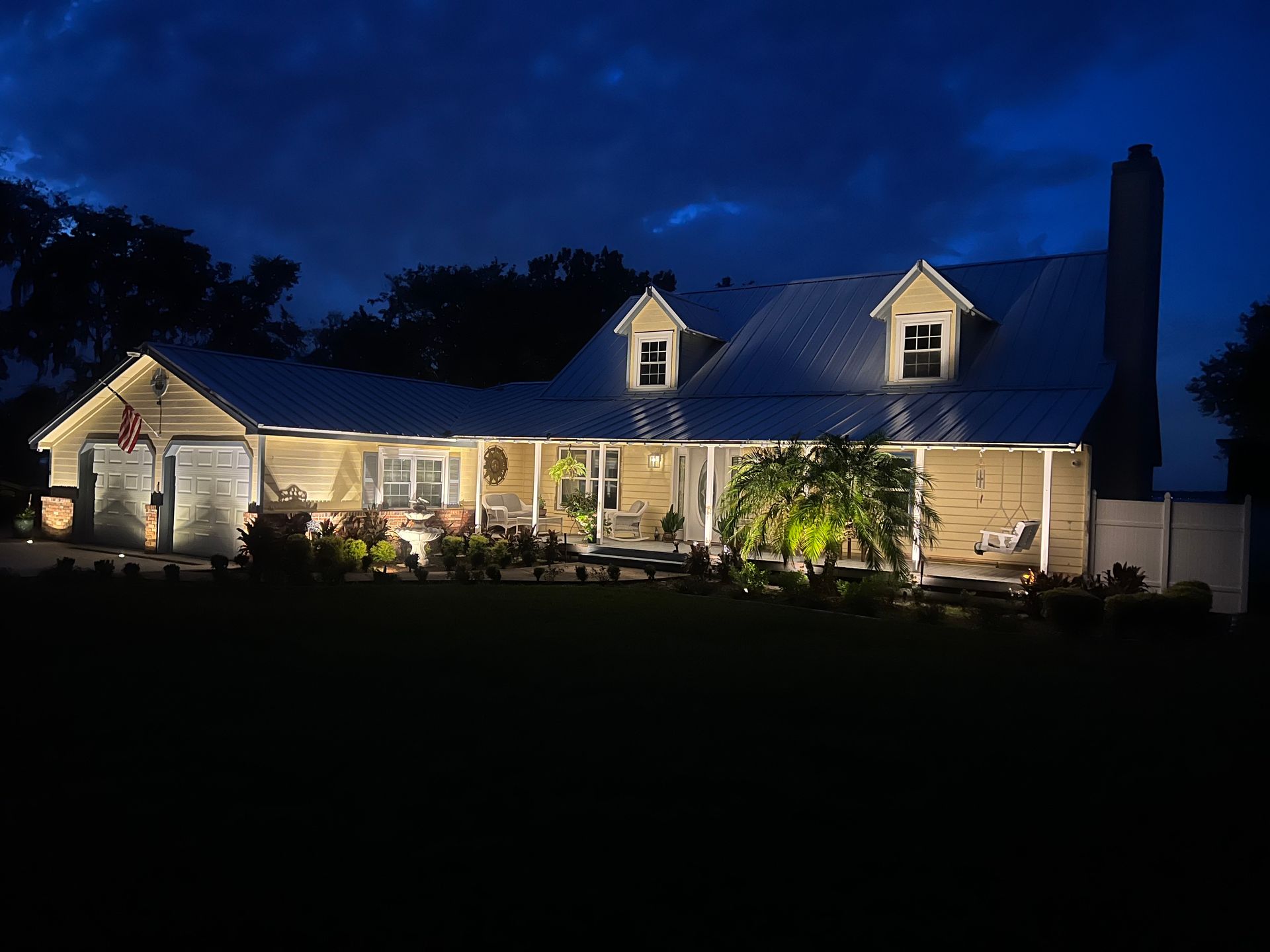 House illuminated at night, with porch lights and landscaping lights, under a dark blue sky.