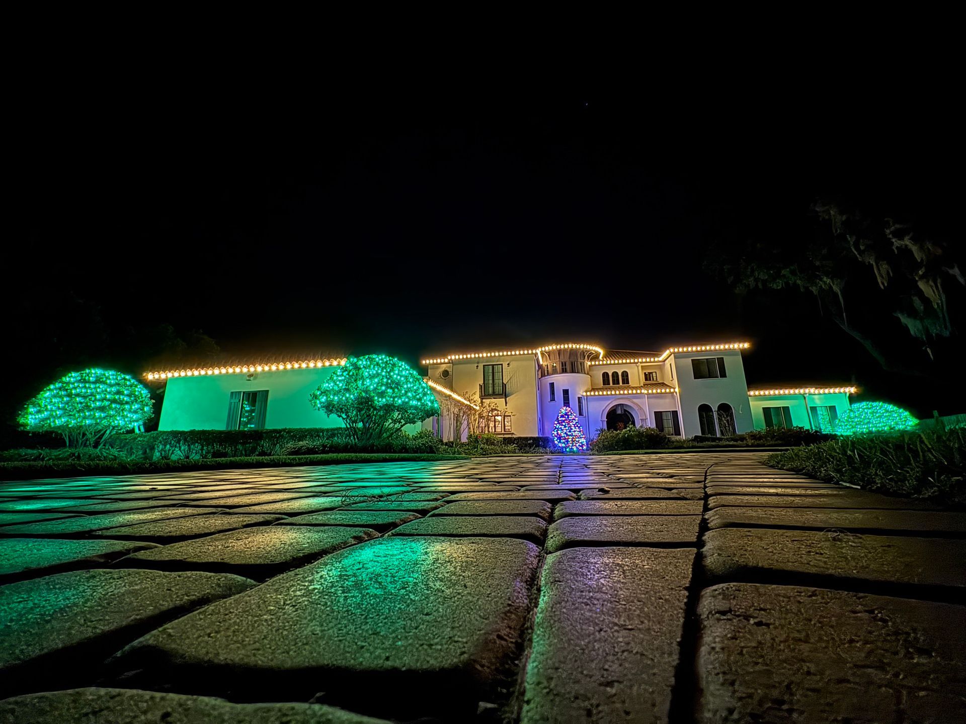 Large house with green and gold Christmas lights, at night; bricks in foreground.