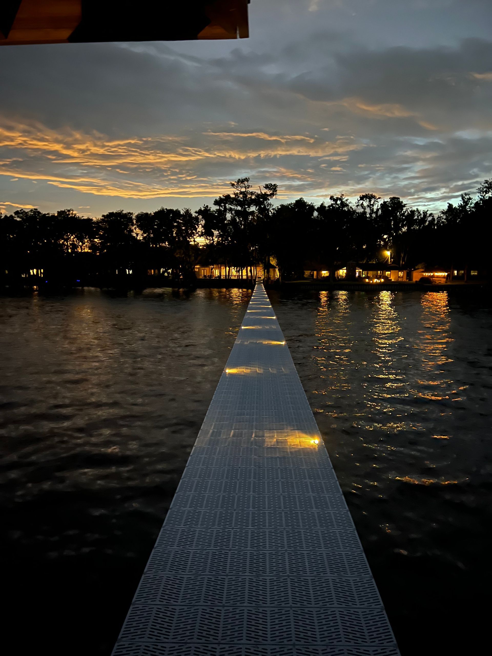 Night scene of illuminated walkway over water, reflecting lights, with dusky sky and trees.