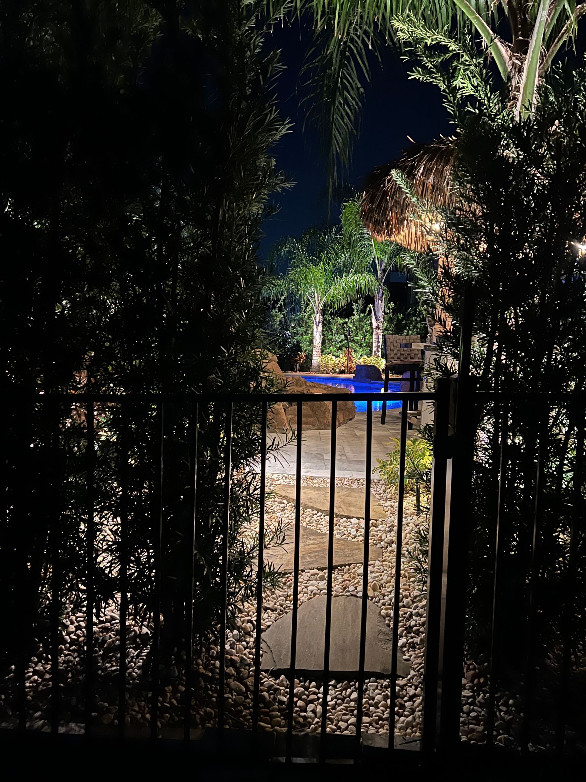 A dark backyard at night. A lit pool can be seen beyond a black fence and gravel path. Trees frame the view.