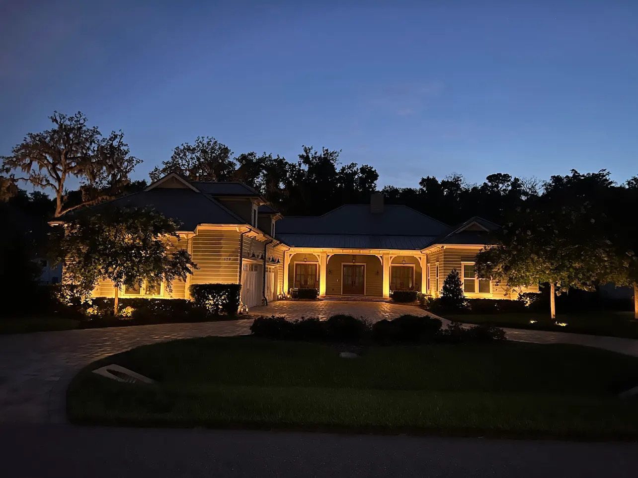 Night view of a house illuminated by outdoor lighting. Trees and a driveway surround the home against a twilight sky.
