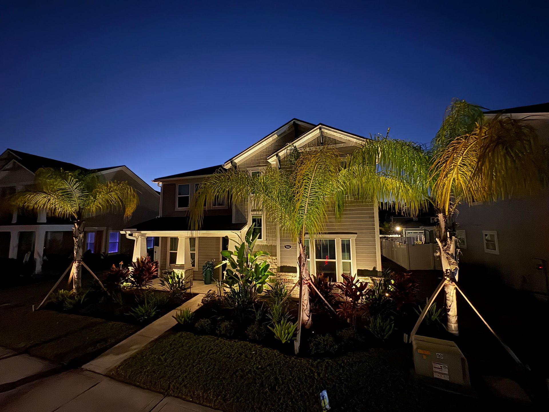 Two-story house with palm trees, lit by outdoor lights at dusk.